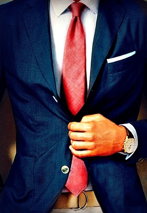 Man in a navy suit and textured red tie adjusting his jacket, showing a white pocket square and wristwatch