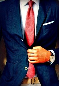 Man in a navy suit and textured red tie adjusting his jacket, showing a white pocket square and wristwatch