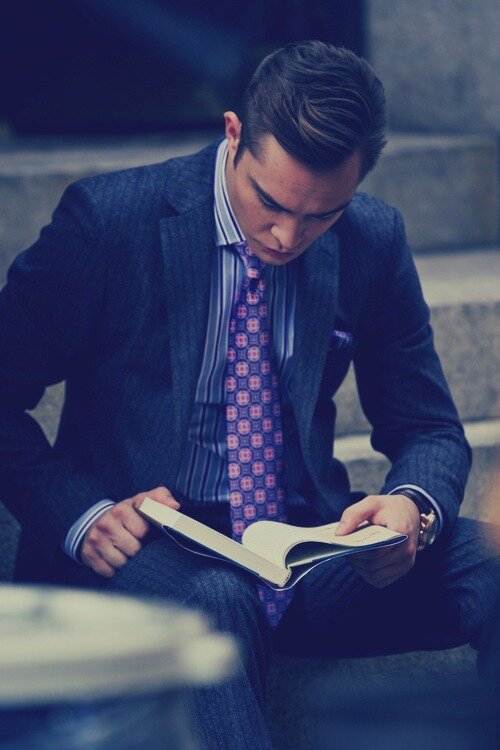 Man in a dark blue suit and purple patterned tie sitting on steps, reading an open book