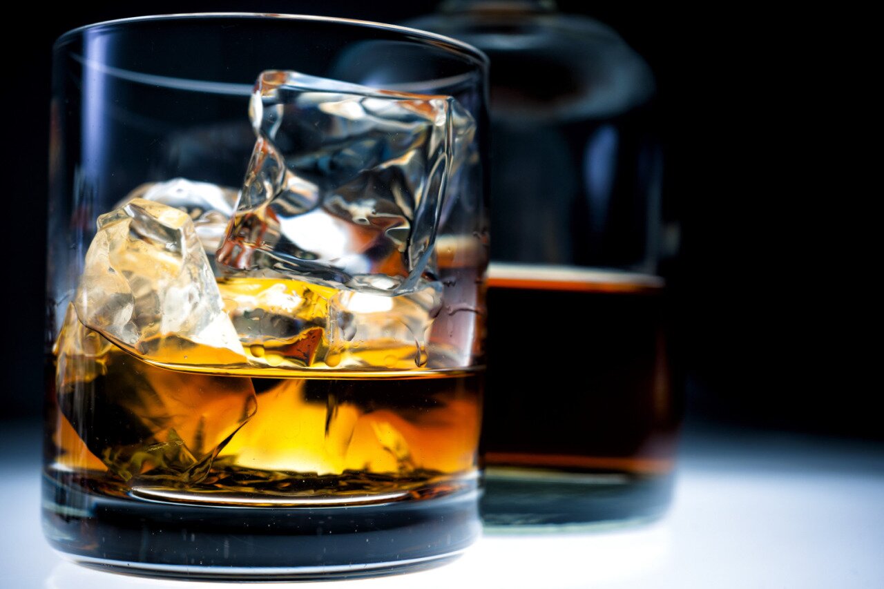 Amber whiskey in a rocks glass with large ice cubes, glowing against a dark bottle behind