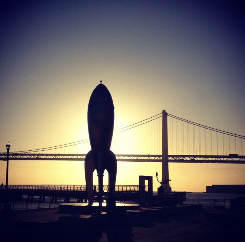 Silhouette of a retro rocket sculpture on a pier with a suspension bridge and golden sunset