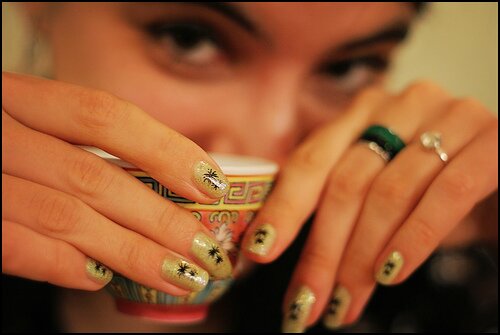 Hands with glitter nails and rings holding an ornate teacup, woman's face softly blurred behind