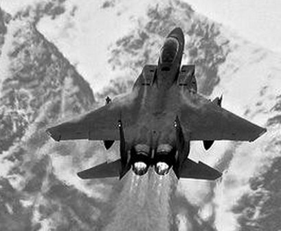 Black-and-white top-down view of an F-15 banking over snowy mountains with bright engine exhaust