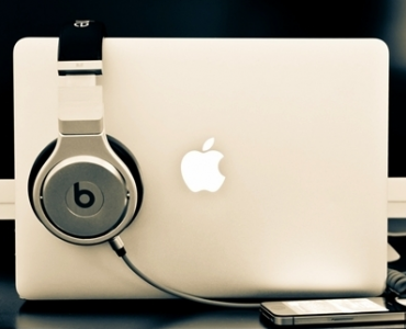 Silver Beats headphones draped over a closed MacBook with glowing Apple logo, cable to a phone