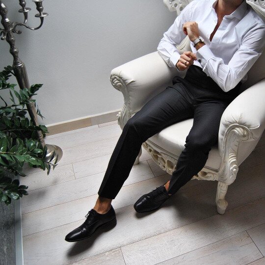 Man in a white shirt and black trousers in an ornate white chair, adjusting a silver cufflink