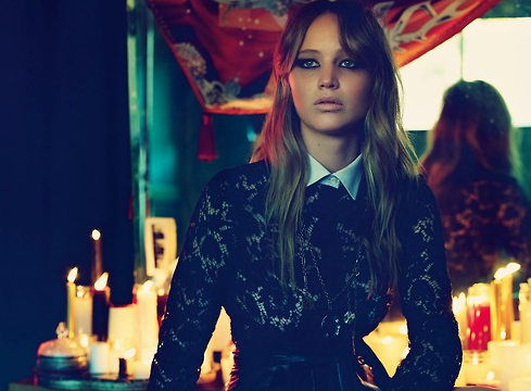 Blonde woman with dark eyeliner in black lace over a white collar, lit by many candles in a dim room