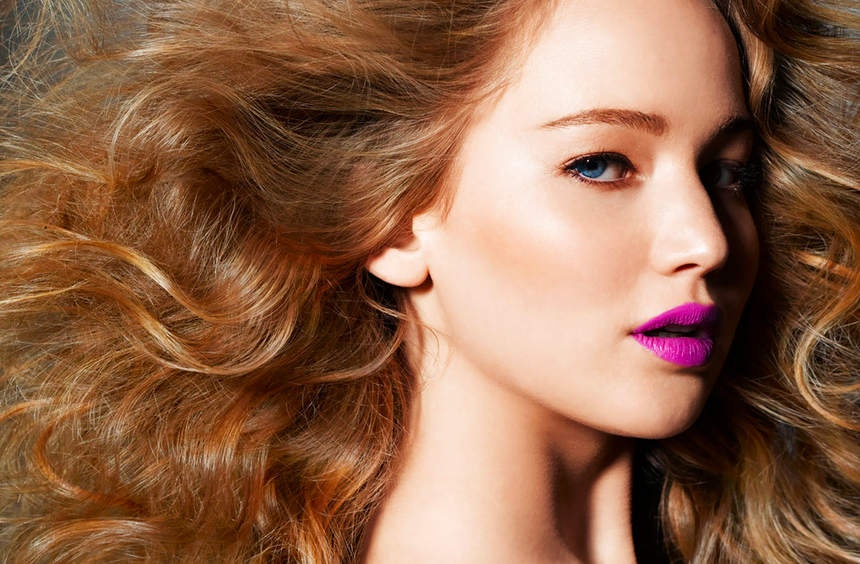Close-up of a blonde woman with voluminous waves looking over her shoulder in bold magenta lipstick
