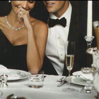 Man in a tuxedo and woman in a black gown smiling at a formal table with champagne and candlelight