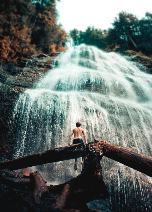 Shirtless man seated on a fallen log looking toward a wide multi-tiered waterfall in a forest