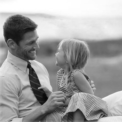 Black-and-white moment of a smiling man in a tie with a young girl in a striped dress outdoors