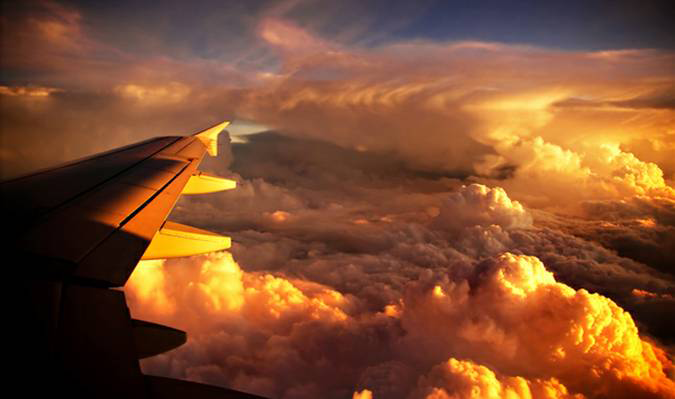 Airplane wing silhouetted above a sea of clouds glowing golden orange at sunset