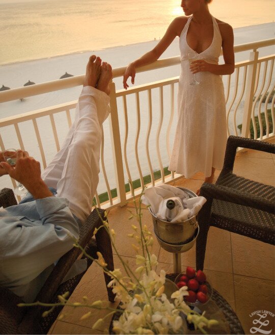 Man with feet on a balcony rail and woman in white dress overlooking a beach sunset with champagne