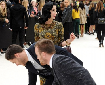 Woman in a mustard floral dress and gold necklace smiling as two suited men bow in the foreground