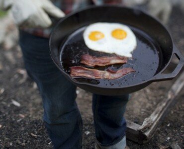 Person in white work gloves holding a cast-iron skillet outdoors with two sunny-side-up eggs and bacon strips arranged like a smiley face