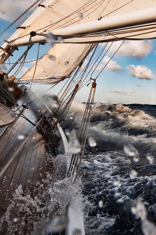 Wooden sailboat heeling hard in rough dark-blue seas, cream sails and spray over the deck