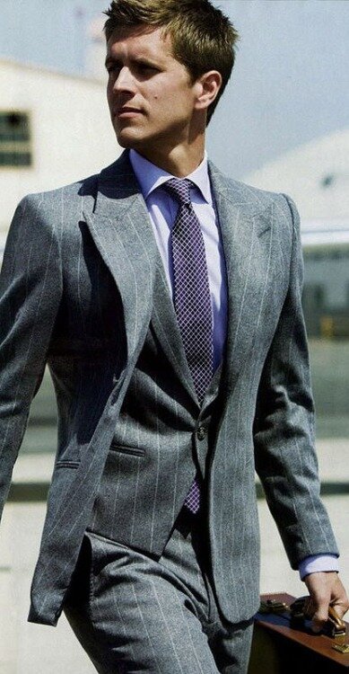 Man in a grey three-piece pinstripe suit and purple tie walking outdoors with a brown briefcase