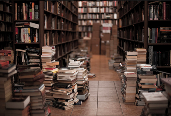 Narrow bookstore aisle lined with packed shelves and tall stacks of books piled on the tiled floor