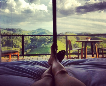 Bare legs crossed on bedsheets facing a balcony view of green mountains under cloudy skies