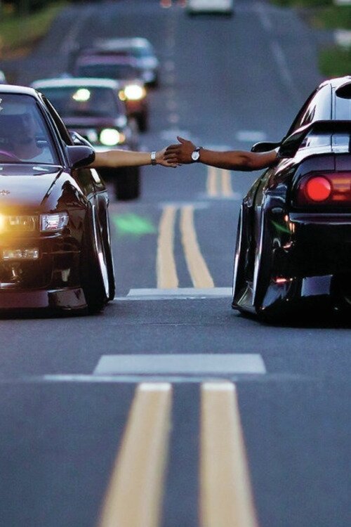 Two low sports cars side by side as drivers' hands reach to shake over the yellow road lines