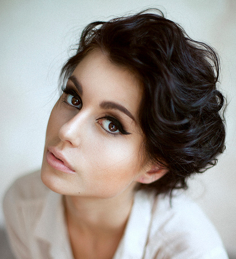 Close-up of a woman with short dark wavy hair and winged eyeliner looking up at the camera