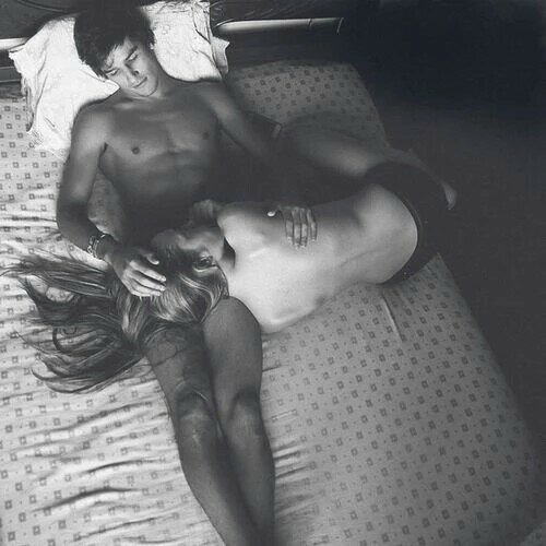 Black-and-white overhead of a shirtless man in bed with a topless woman resting across his lap