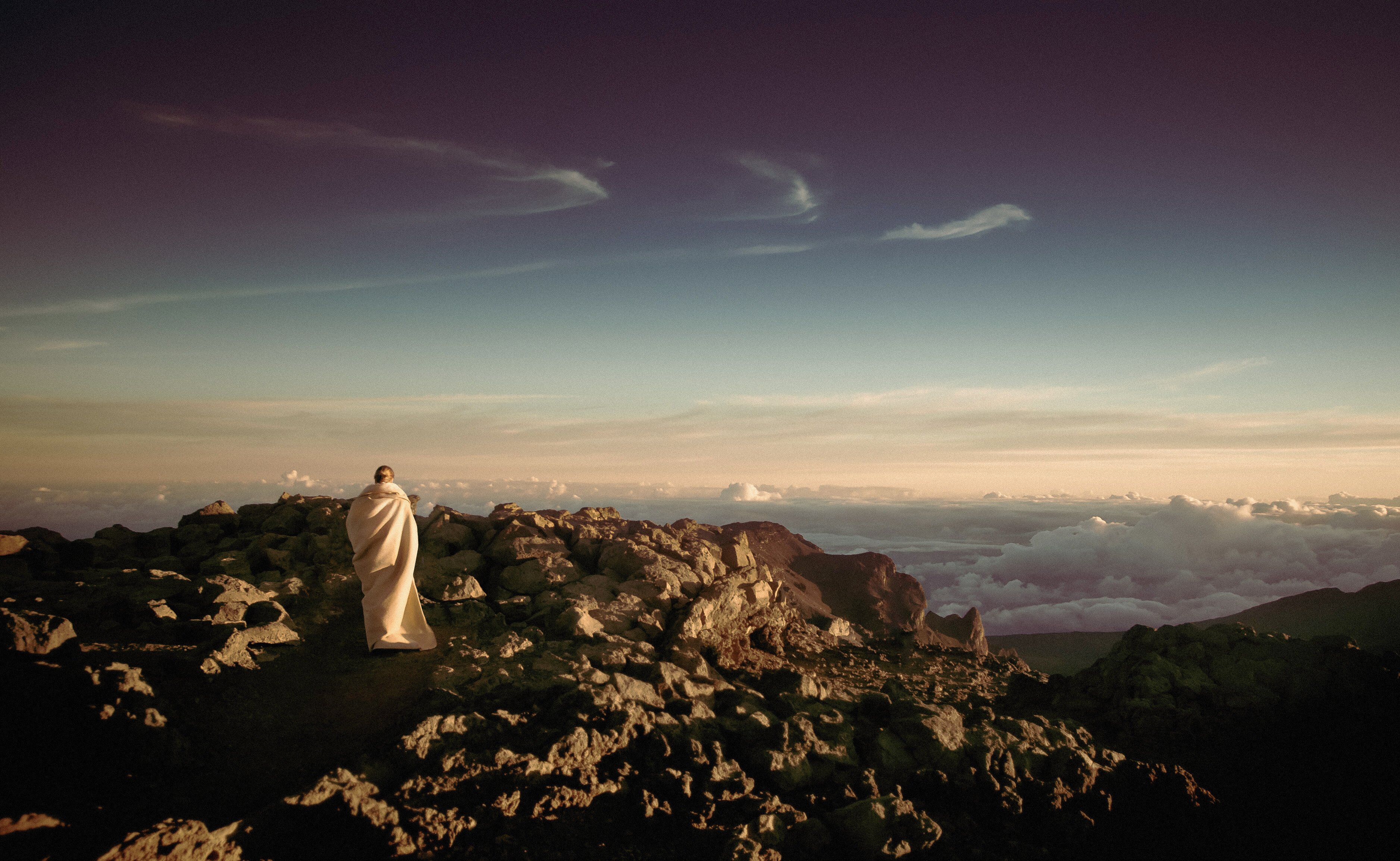 Person wrapped in a beige blanket on a rocky summit overlooking a sea of clouds at dawn