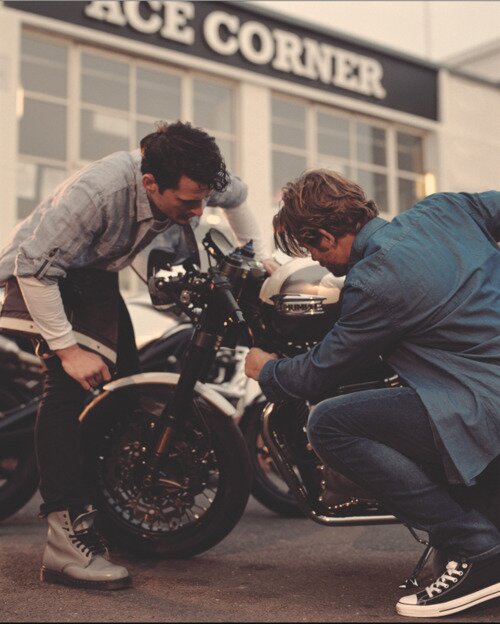 Two men examine a black Triumph motorcycle outside a shop with an Ace Corner sign