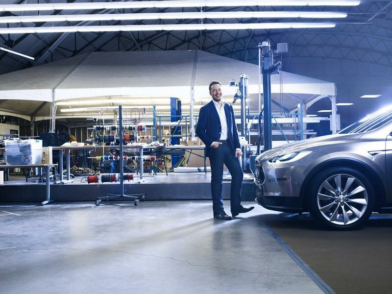 Elon Musk smiling beside a silver Tesla in a brightly lit industrial workshop