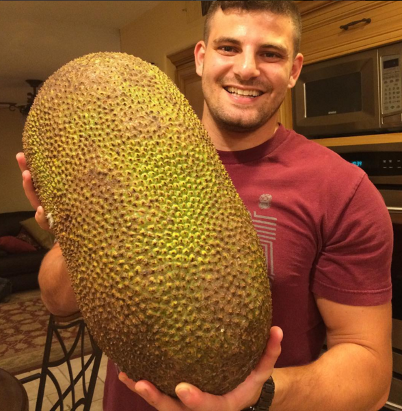 Smiling man in a maroon shirt holding a huge spiky green jackfruit nearly as large as his torso