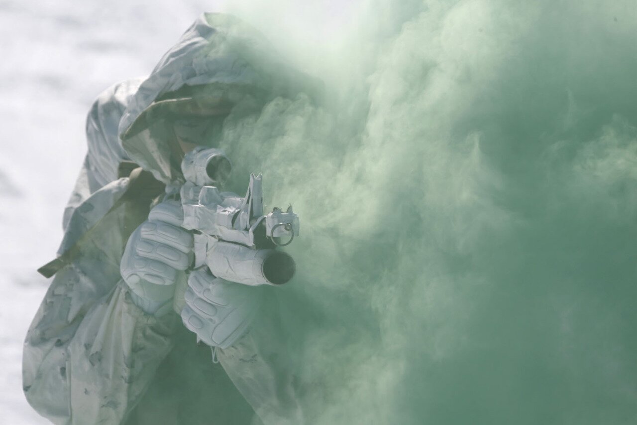 Soldier in white arctic camouflage aiming a scoped rifle amid pale green smoke in snow