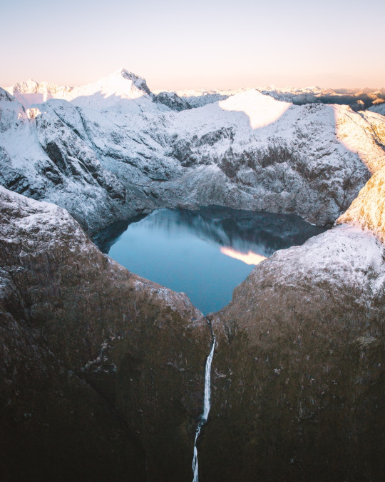 Snowy peaks around a still blue alpine lake spilling into a thin waterfall down a dark cliff