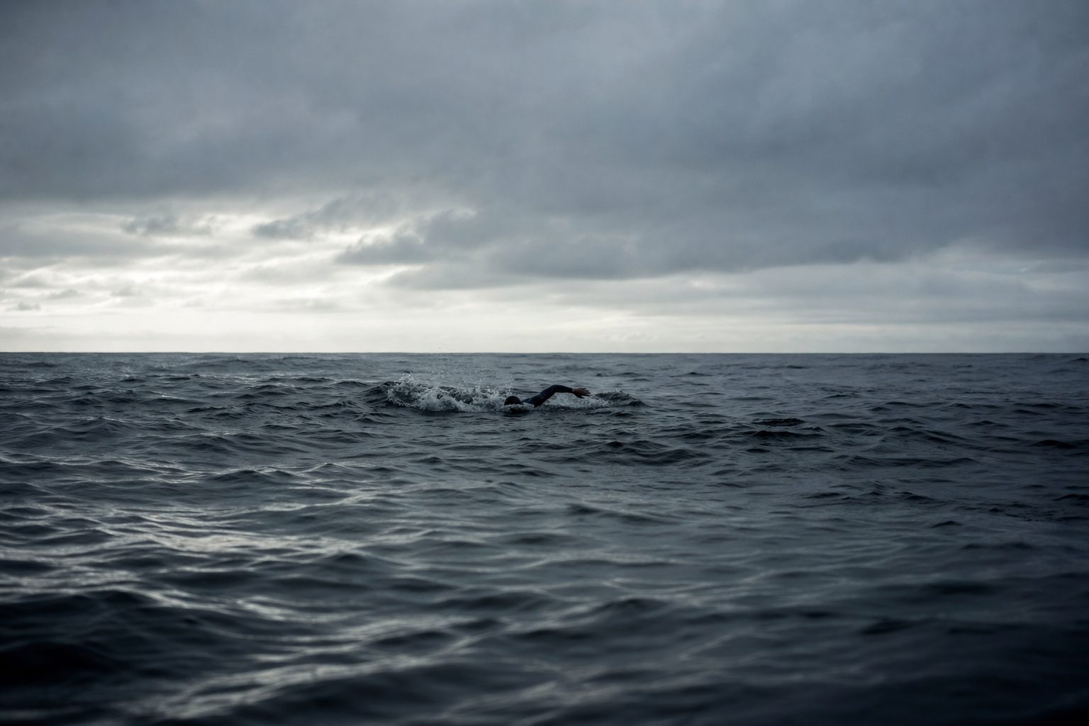 Vast open ocean under an overcast sky with a distant lone swimmer on the horizon