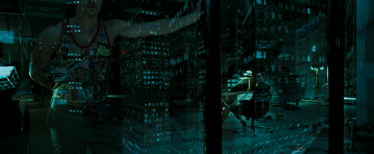 Teal-toned shot of a man in a graphic tank by glass reflecting a city skyline, another man at a desk