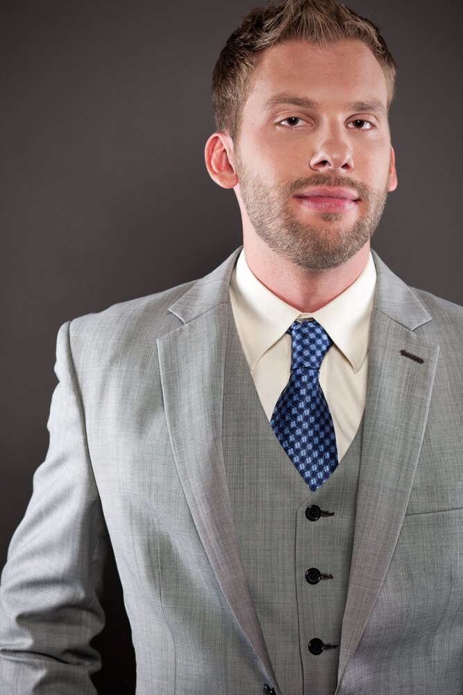 Bearded man in a light grey three-piece suit and blue patterned tie smiling against a dark backdrop
