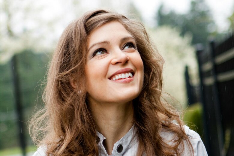 Young woman with long wavy brown hair smiling and looking upward outdoors against blurred greenery and white blossoms