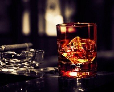 Amber whiskey with a large ice cube beside a lit cigar in a glass ashtray on a dark bar top