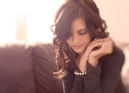Woman with wavy brown hair looking down serenely, hands clasped near her face, pearl bracelet