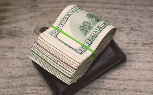 Thick stack of hundred-dollar bills bound with a green rubber band atop a brown Coach wallet