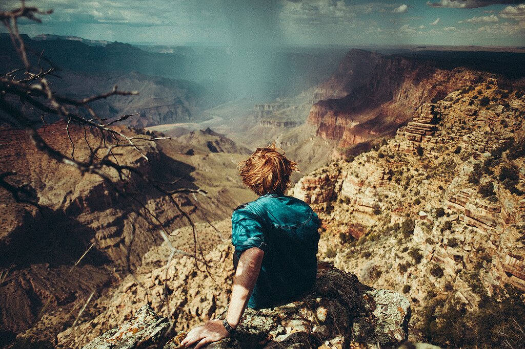 Man with messy red hair sits on a rocky ledge overlooking a vast canyon and distant rainstorm