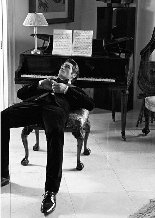 Black-and-white shot of a man in a velvet tuxedo lounging on a piano bench beside a grand piano