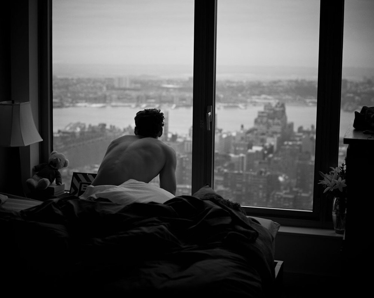 Black-and-white photo of a shirtless man sitting on a bed facing a floor-to-ceiling window overlooking a hazy city skyline