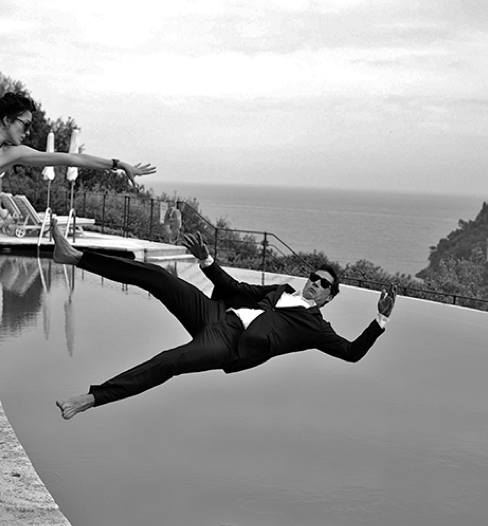 Man in a dark suit and sunglasses falling backward mid-air into an oceanfront infinity pool