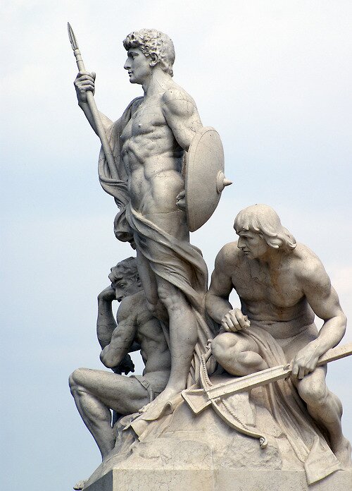 Marble sculpture of three muscular men with spear, shield, and crossbow against an overcast sky
