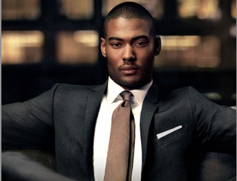 Black man in a dark grey suit, tan tie, and white pocket square seated confidently against warm bokeh lights