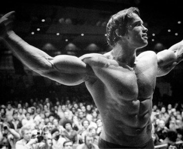 Black-and-white bodybuilder with arms wide posing on stage before a packed arena audience