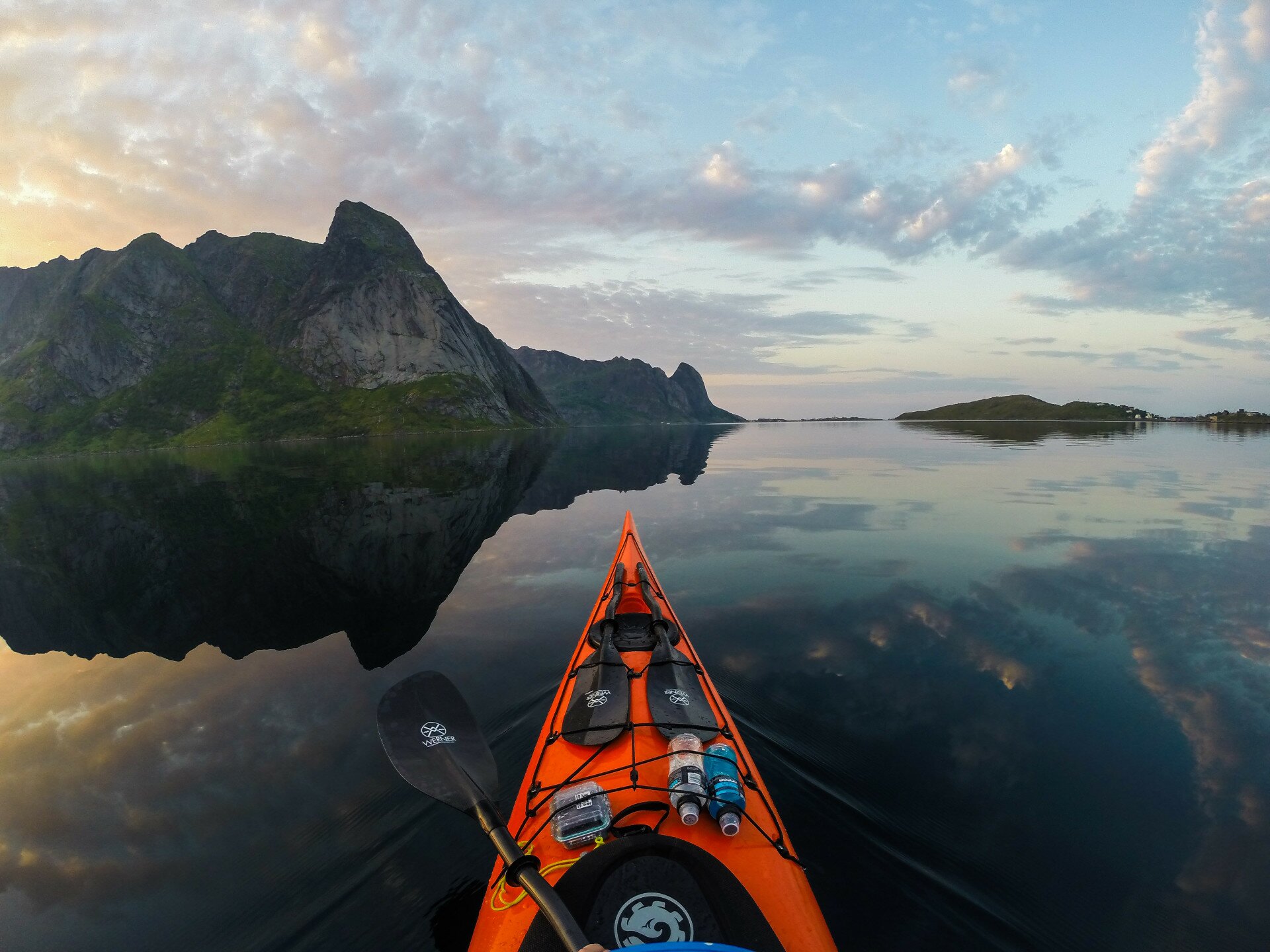 POV from an orange kayak on glassy water reflecting dark mountains under a soft golden sky
