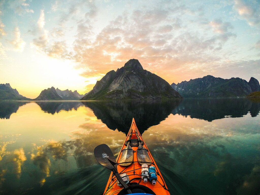 Orange kayak bow aimed at a towering triangular peak mirrored in perfectly still fjord water