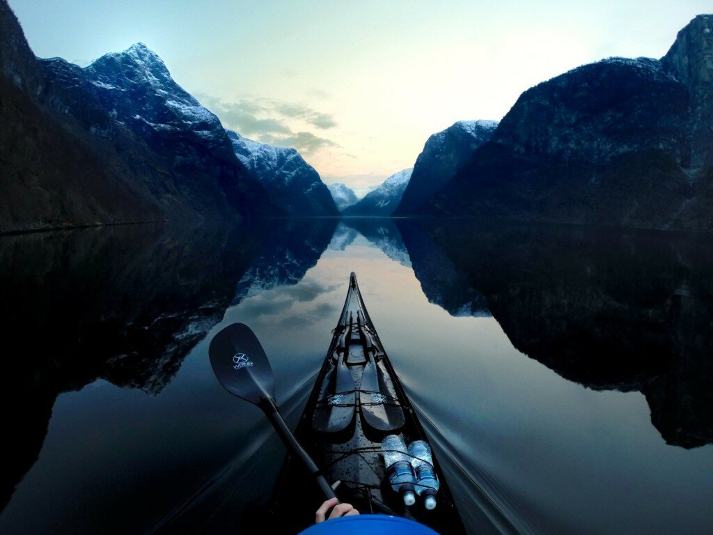 Black kayak bow on a mirror-still fjord framed by steep snow-dusted mountains on both sides