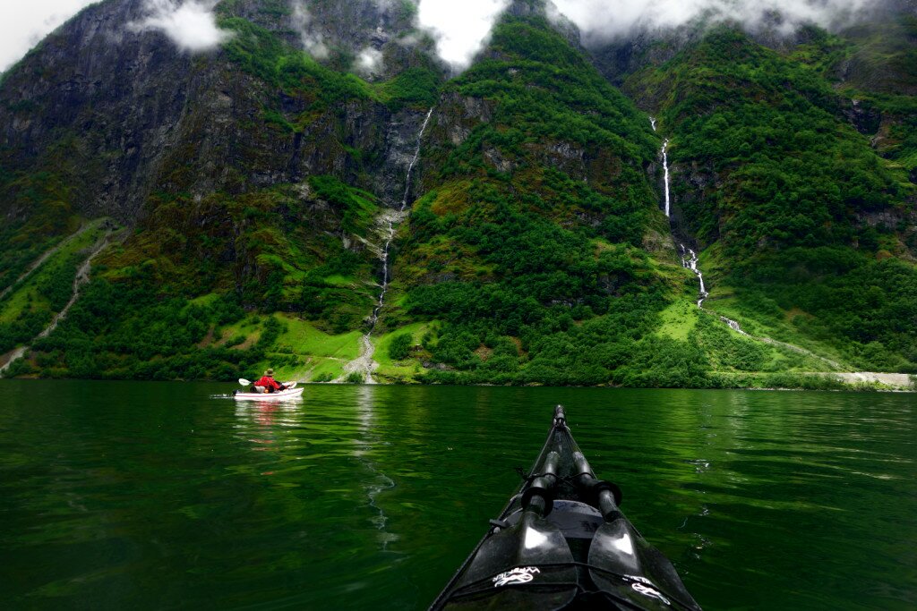 Black kayak view of a red-jacket paddler ahead of a green cliff laced with thin waterfalls