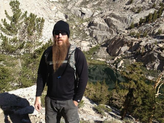 Long-bearded hiker in beanie and sunglasses before a green alpine lake and rocky peaks